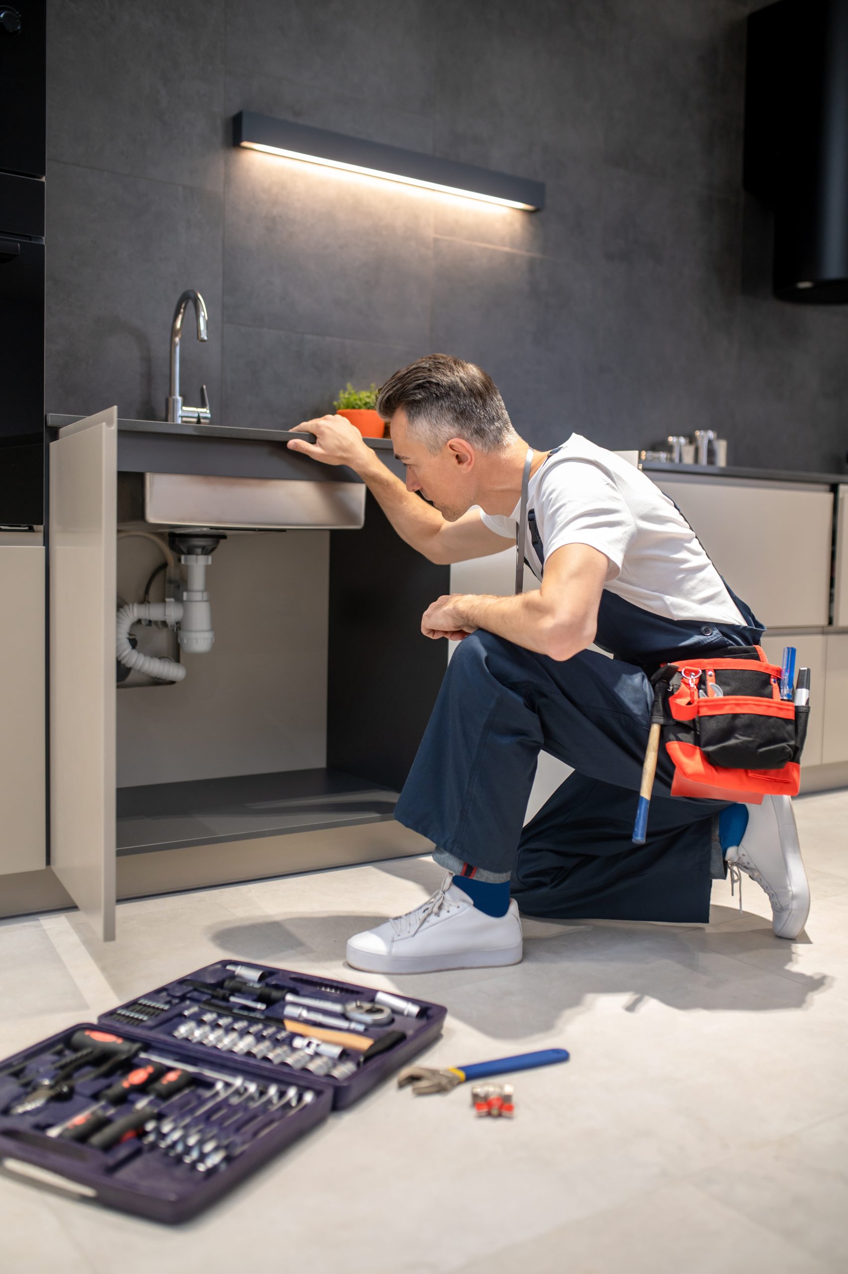 man kneeling down touching sink inspecting pipes scaled